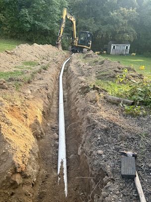 An excavator digs a trench for a long white pipe on a grassy landscape.