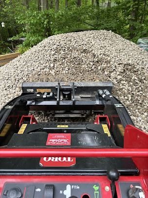 A Toro skid steer loader with a bucket full of gravel outdoors, in a wooded setting.