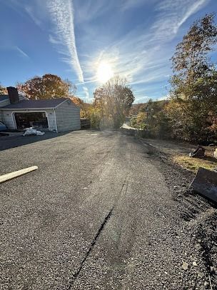Gravel driveway on a sunny day, a house with a garage on the left, trees and blue sky in the background.