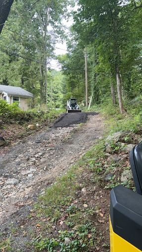 A gravel road being paved by a bobcat, leading uphill between trees toward a house.