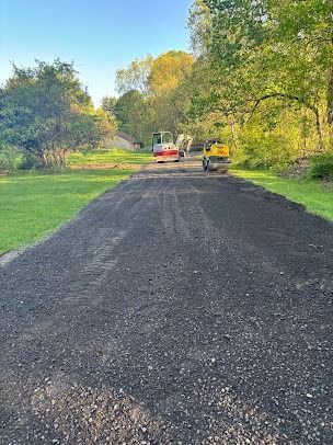 Black asphalt driveway with two construction vehicles, surrounded by green grass and trees.