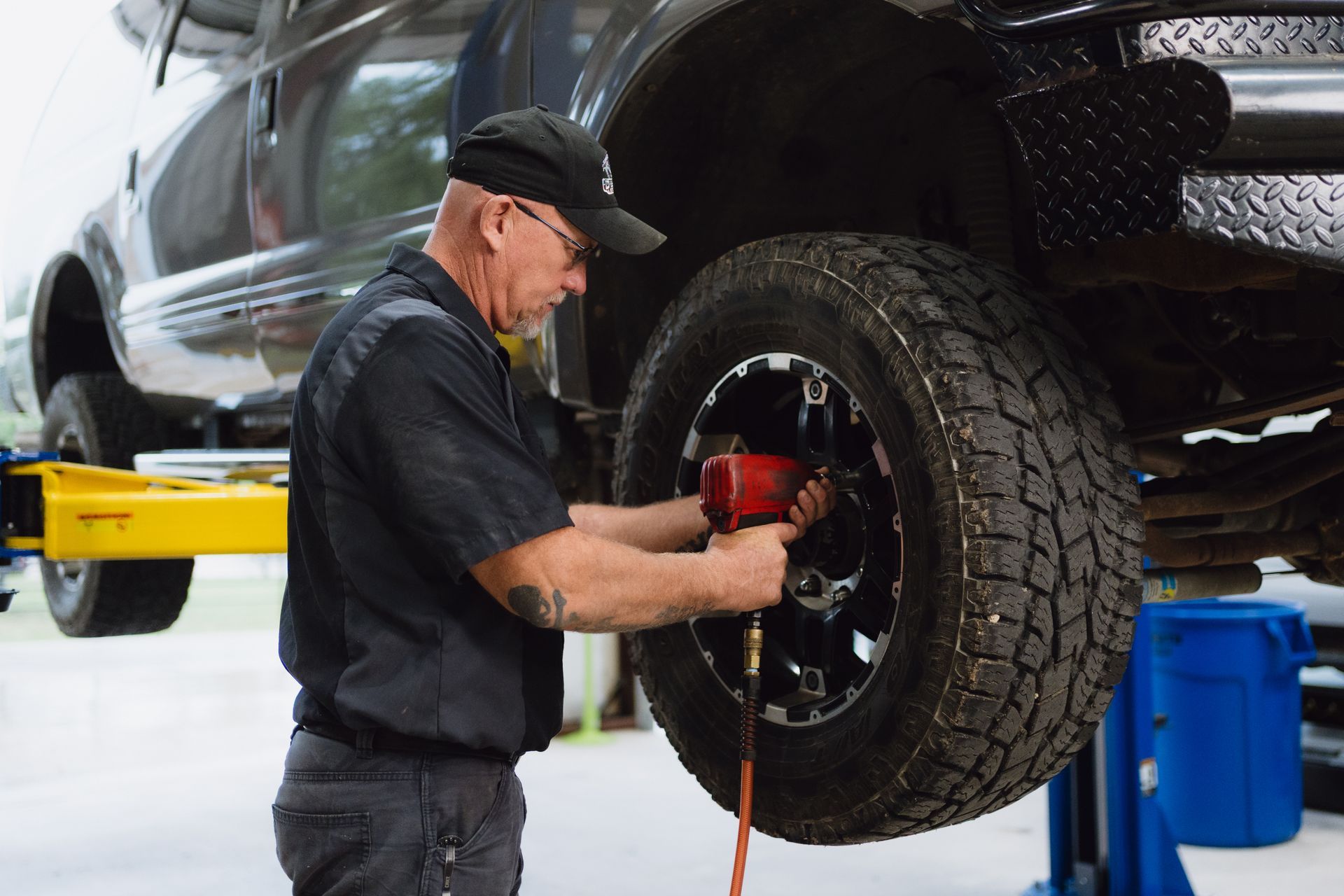 Mechanic using a red power tool to tighten a wheel on a raised black vehicle in a garage | Texas Hill Country Diesel and Automotive
