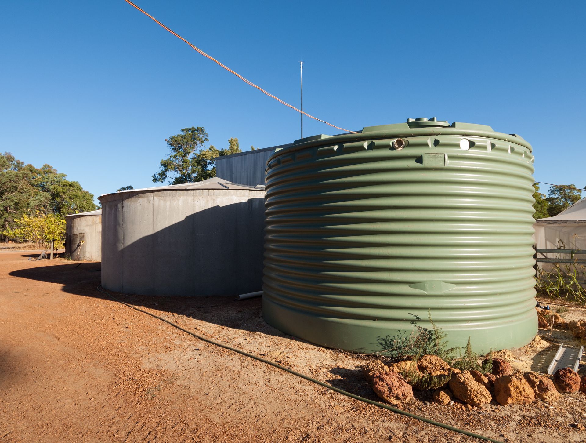 Large green and grey corrugated water tanks sitting on red dirt in a sunny, rural landscape.