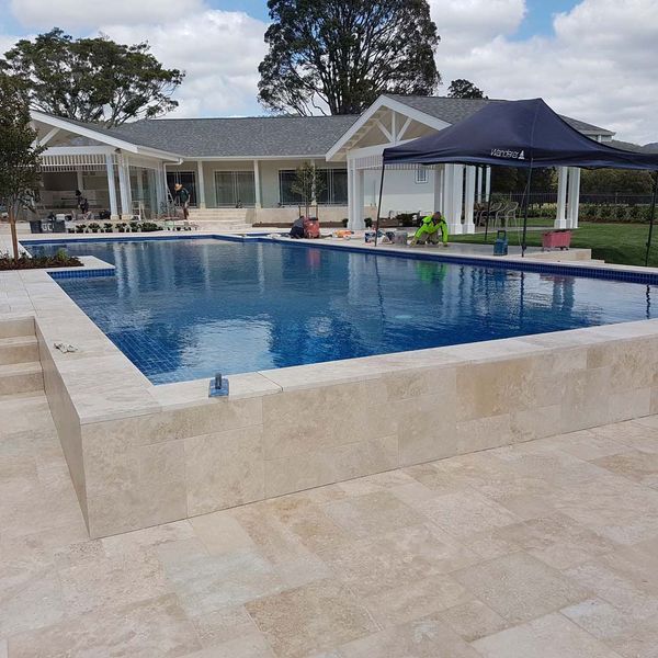 A raised, rectangular swimming pool with blue water and beige stone coping, set on a patio in front of a modern house.