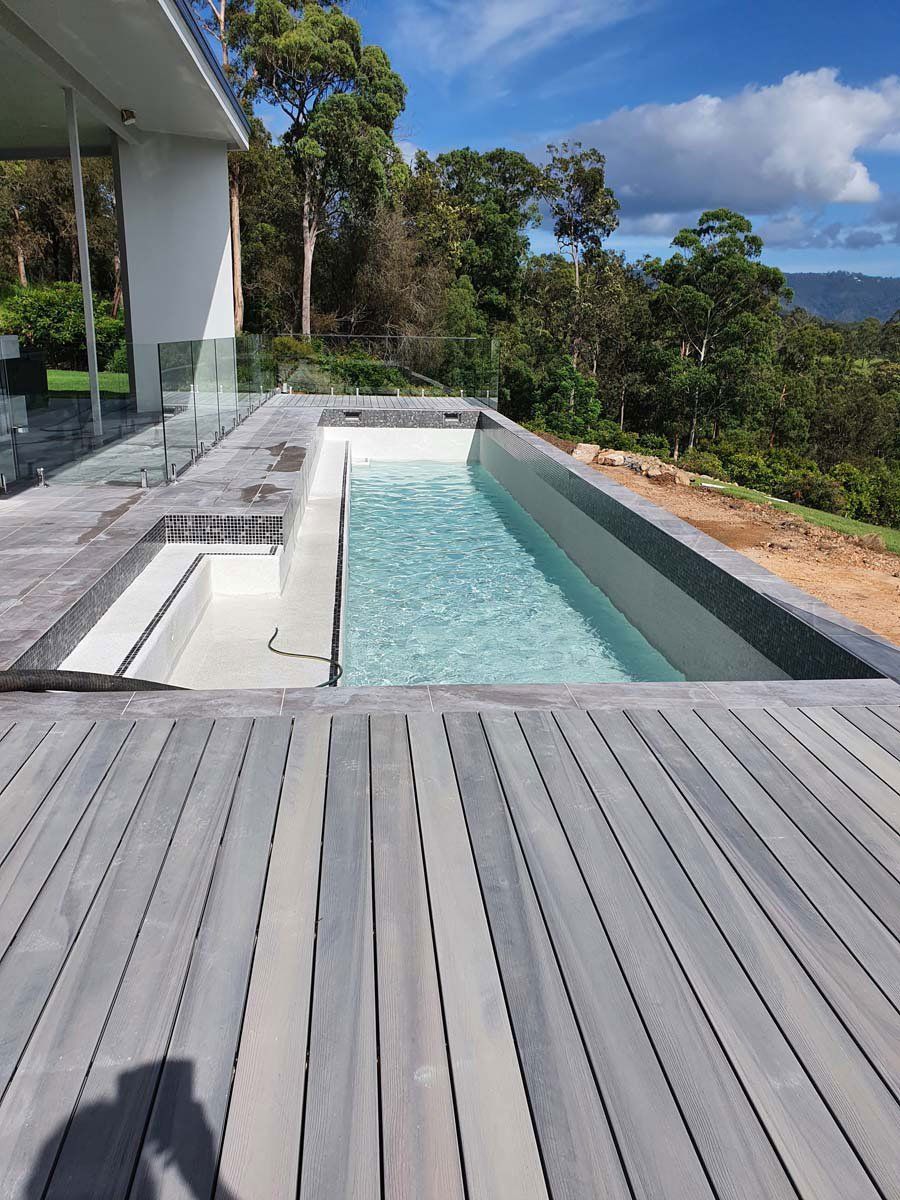 A modern infinity pool with gray decking sits on a hilltop terrace overlooking a lush, forested valley under a blue sky.