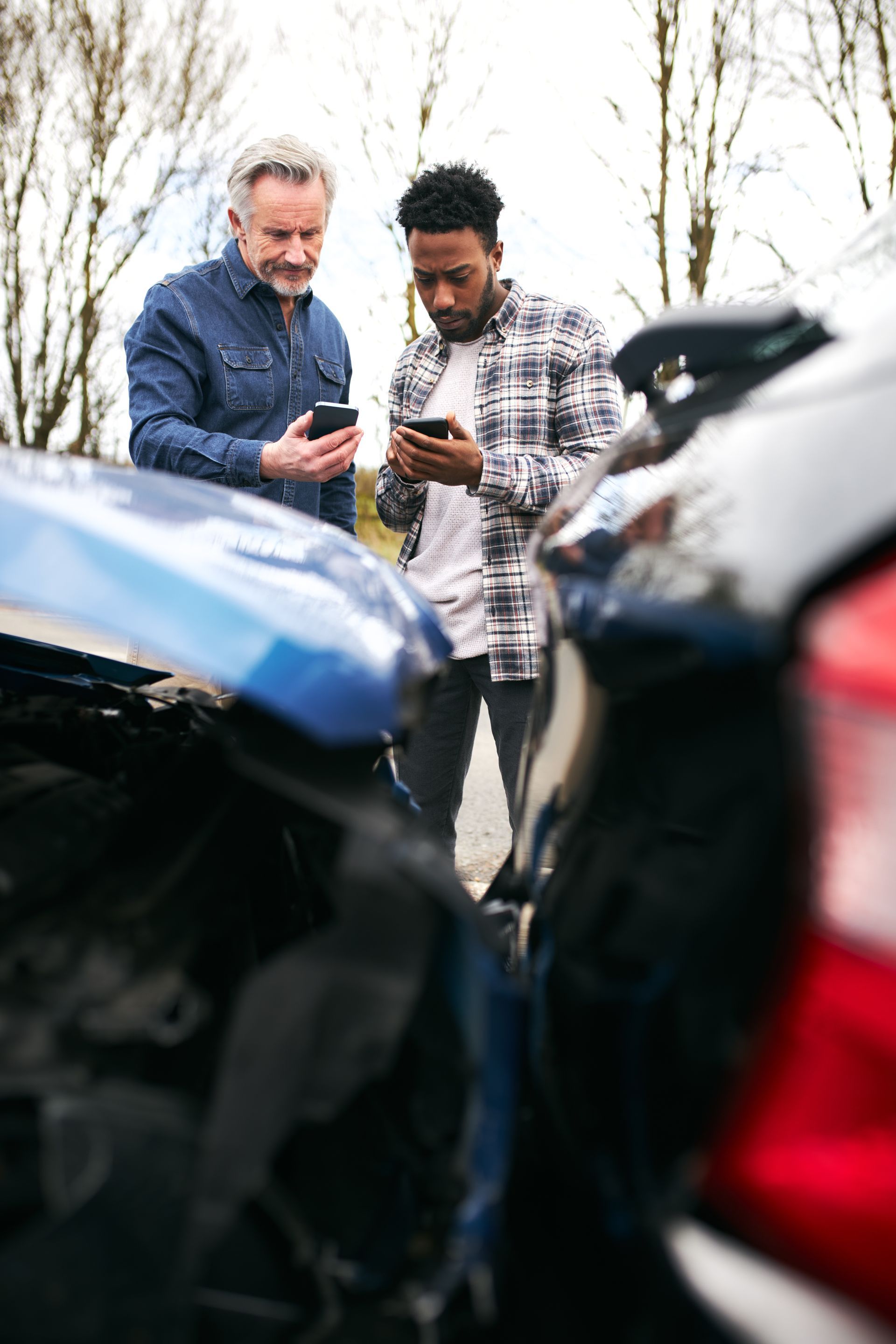 Two men are looking at their phones in front of a car accident.