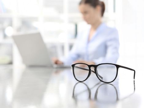 Un paio di occhiali è appoggiato su un tavolo davanti a una donna che sta usando un computer portatile.