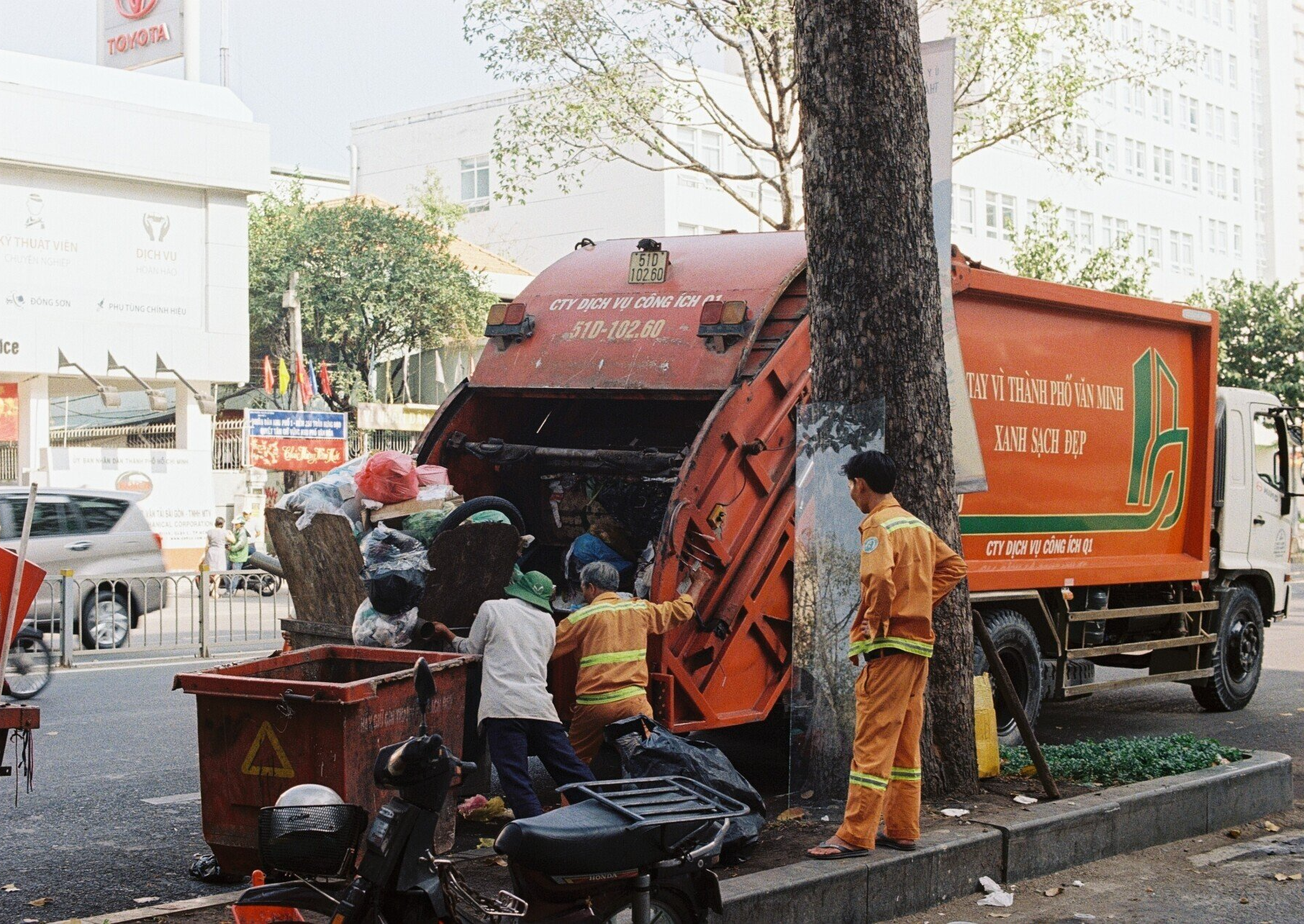 An orange trash truck with three men piling bags of garbage into the back of the truck