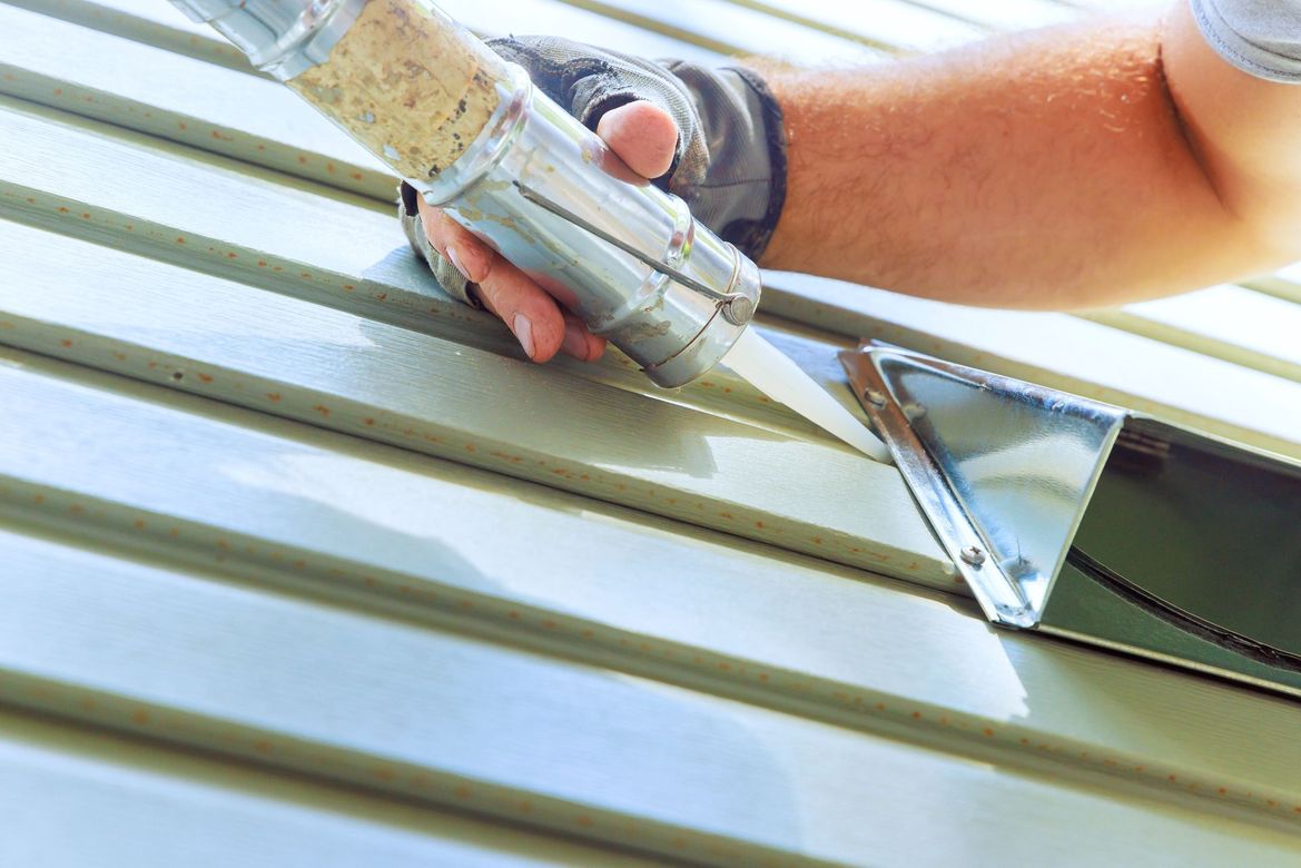 Une personne calfeutre un revêtement métallique avec un pistolet à calfeutrer à l'extérieur.