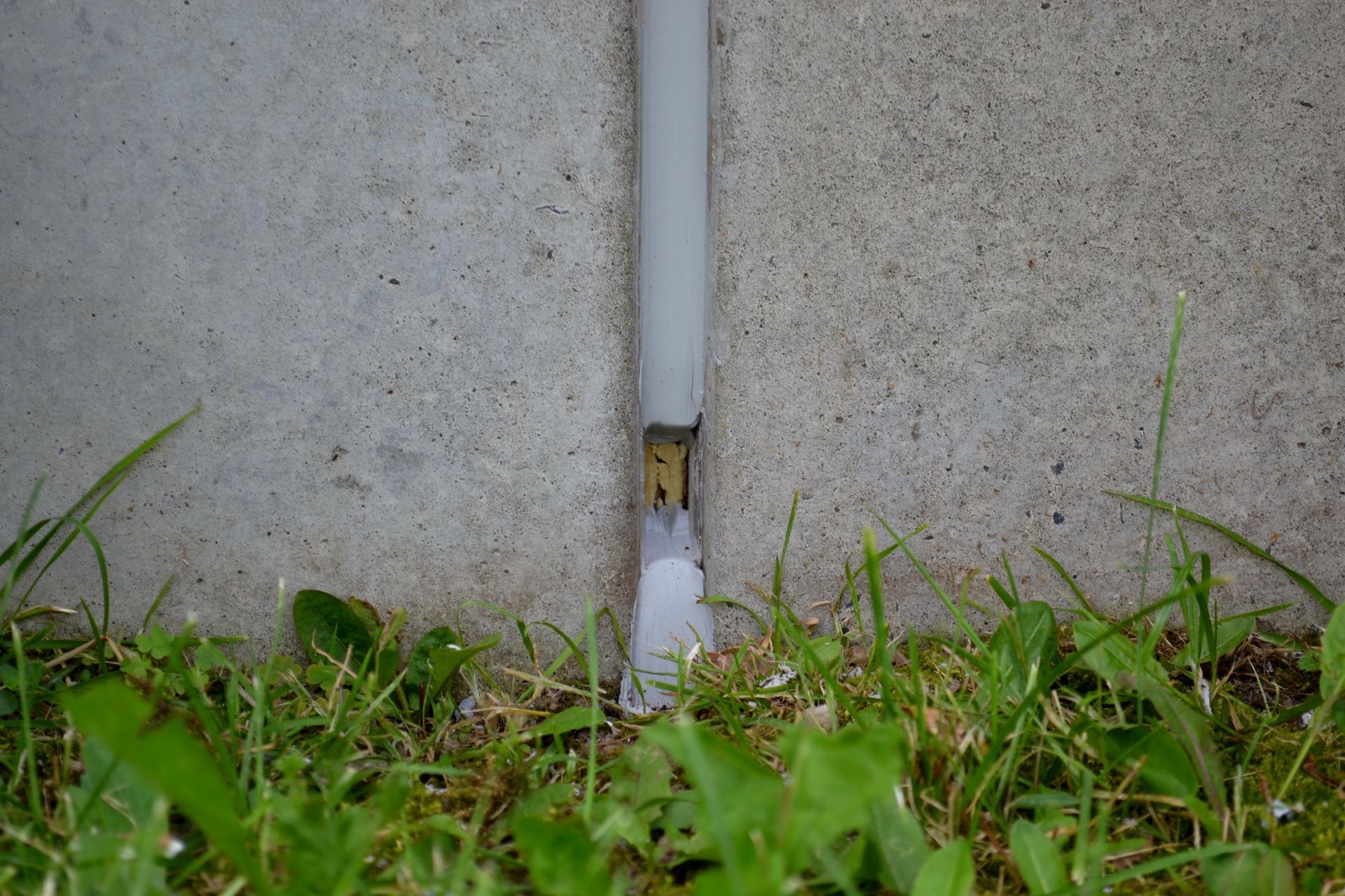 Un conduit métallique longeant un mur en béton, partiellement recouvert, avec de l'herbe verte à la base.