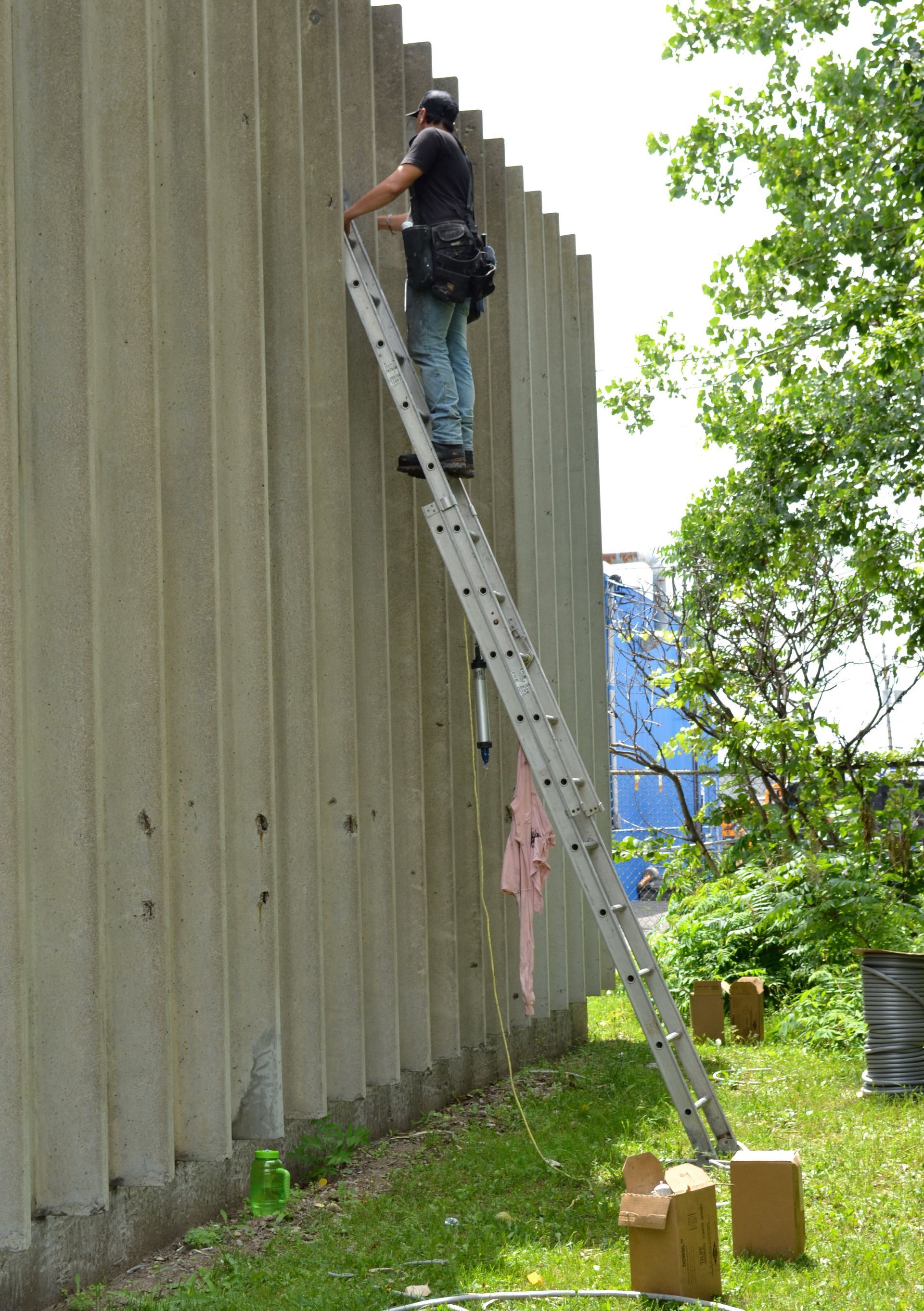 Une personne sur une grande échelle travaille sur un mur en béton nervuré. Herbe verte et arbres en arrière-plan.