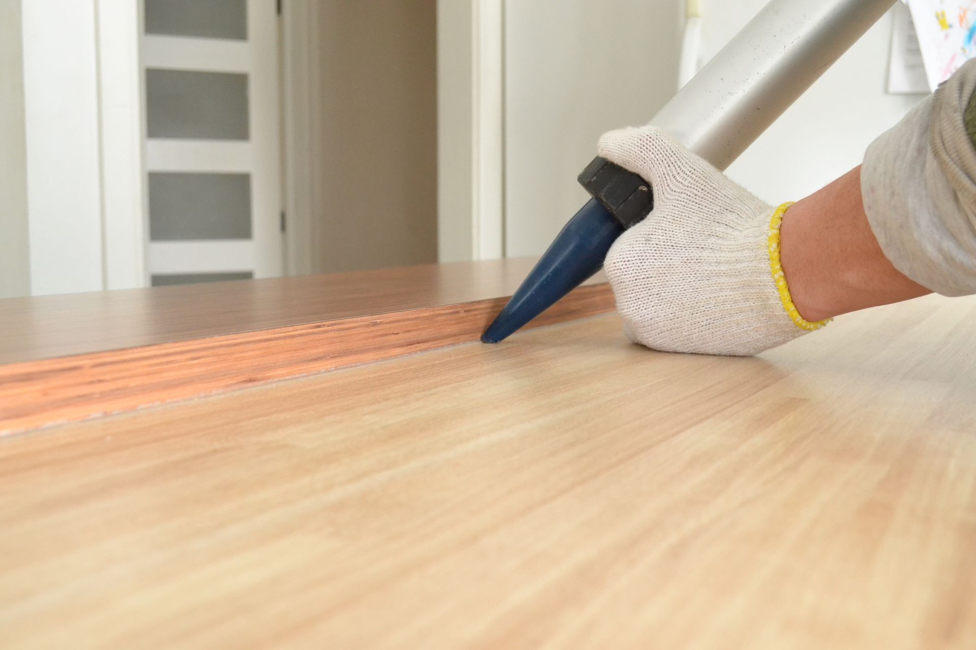 Une personne portant un gant de travail applique du calfeutrage sur une surface en bois avec un pistolet à calfeutrer.