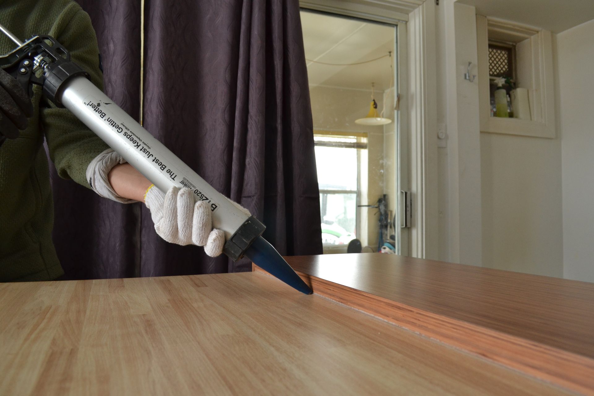 Une personne portant des gants utilise un pistolet à calfeutrer pour appliquer du mastic sur une surface en bois près d'une porte et d'une fenêtre.