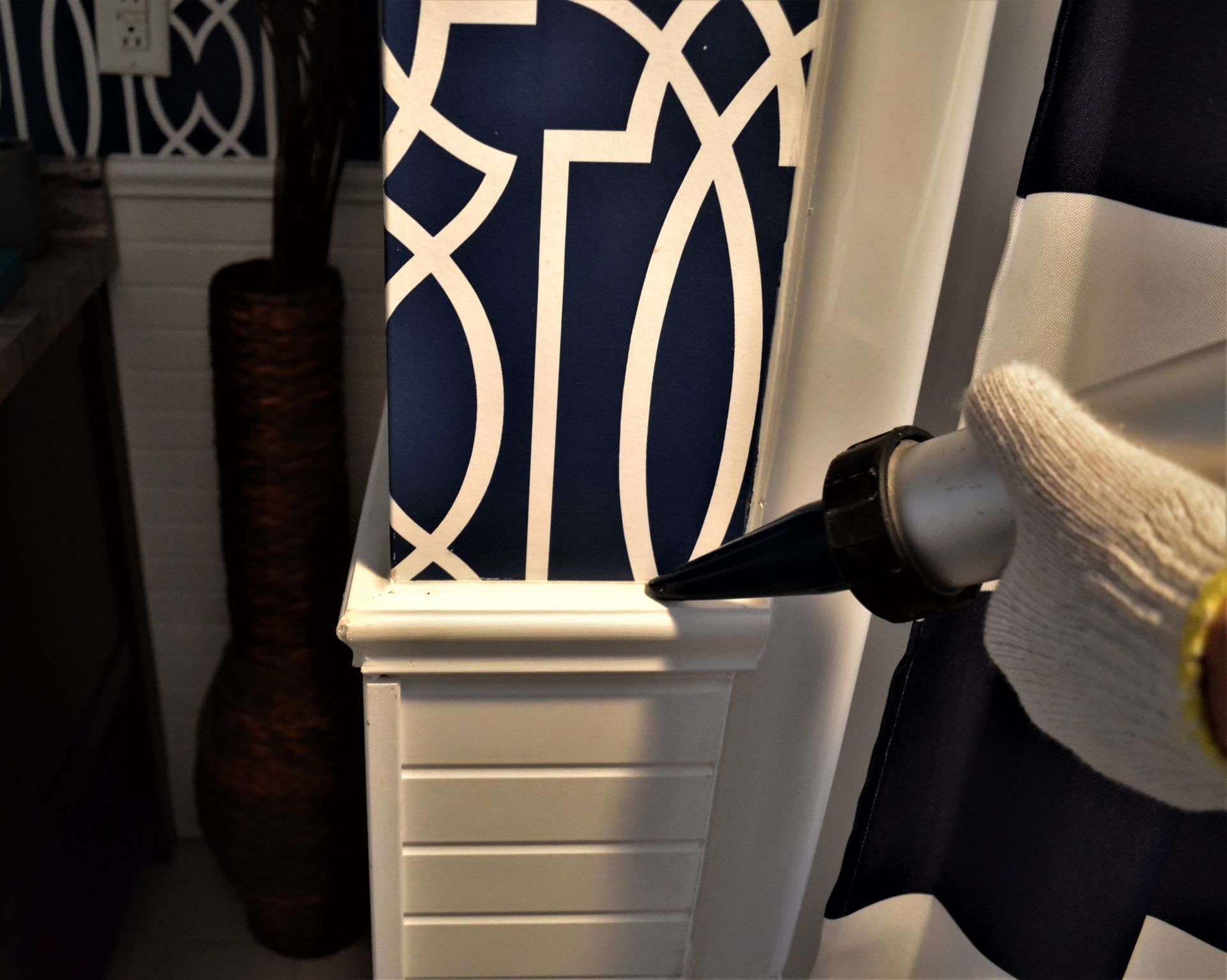 Application de calfeutrage sur une garniture blanche autour d'une colonne à motifs bleus et blancs, une main gantée tient un pistolet à calfeutrer.