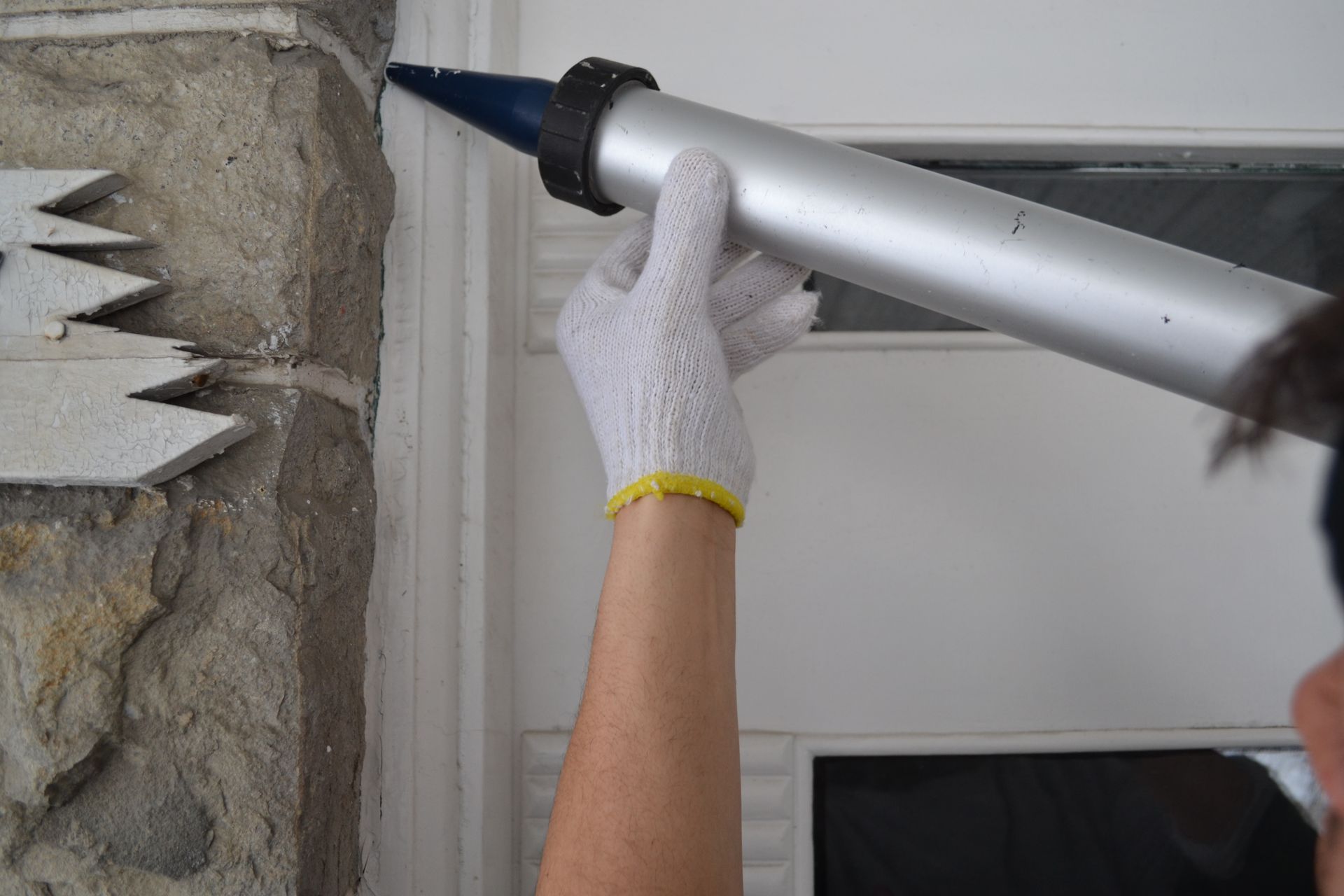 Une personne calfeutrant une garniture blanche autour d'un cadre de porte avec un pistolet à calfeutrer, portant des gants.
