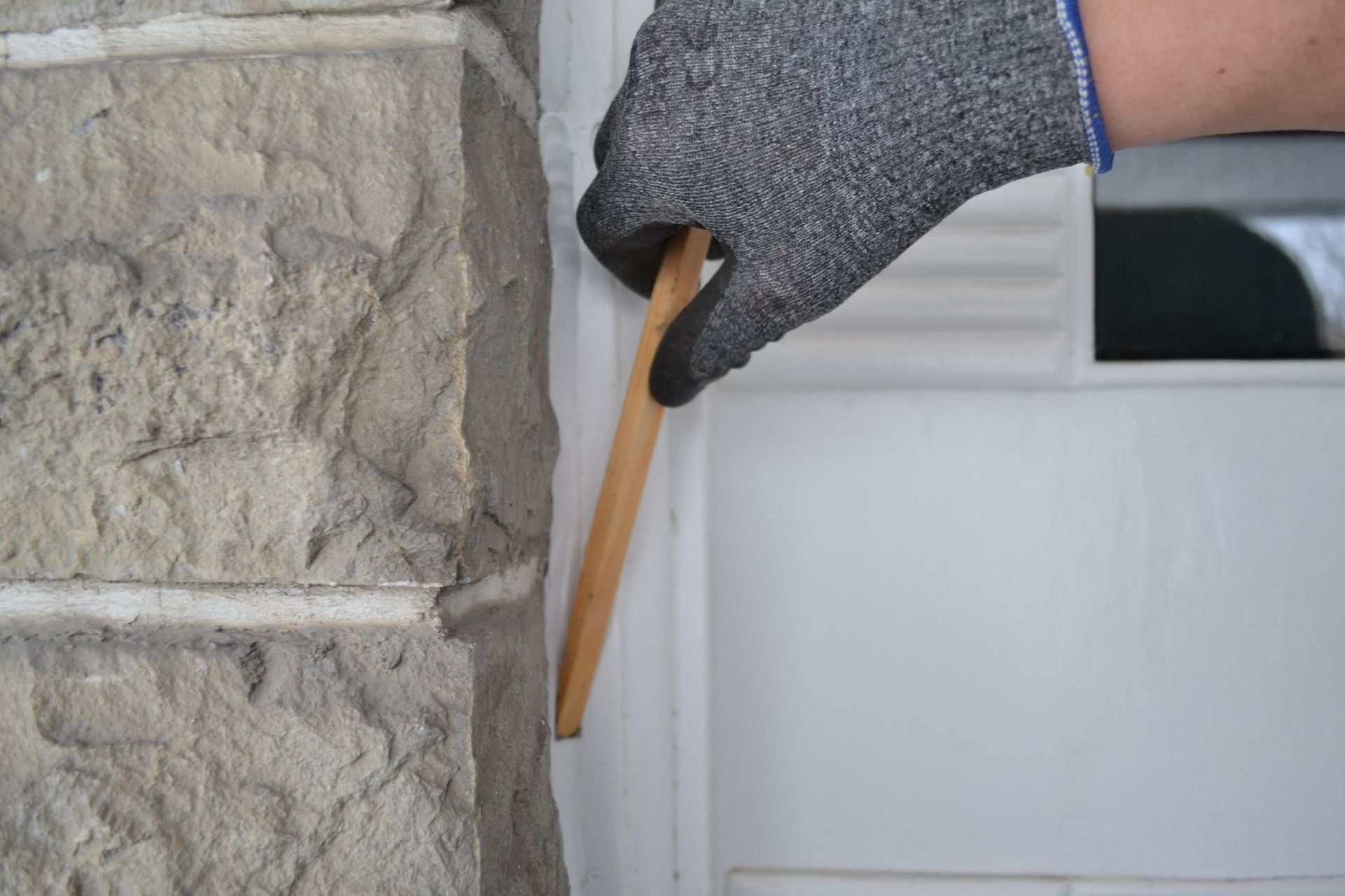 Main gantée utilisant un outil en bois pour retirer quelque chose d'un cadre de porte blanc à côté d'un mur de pierre.