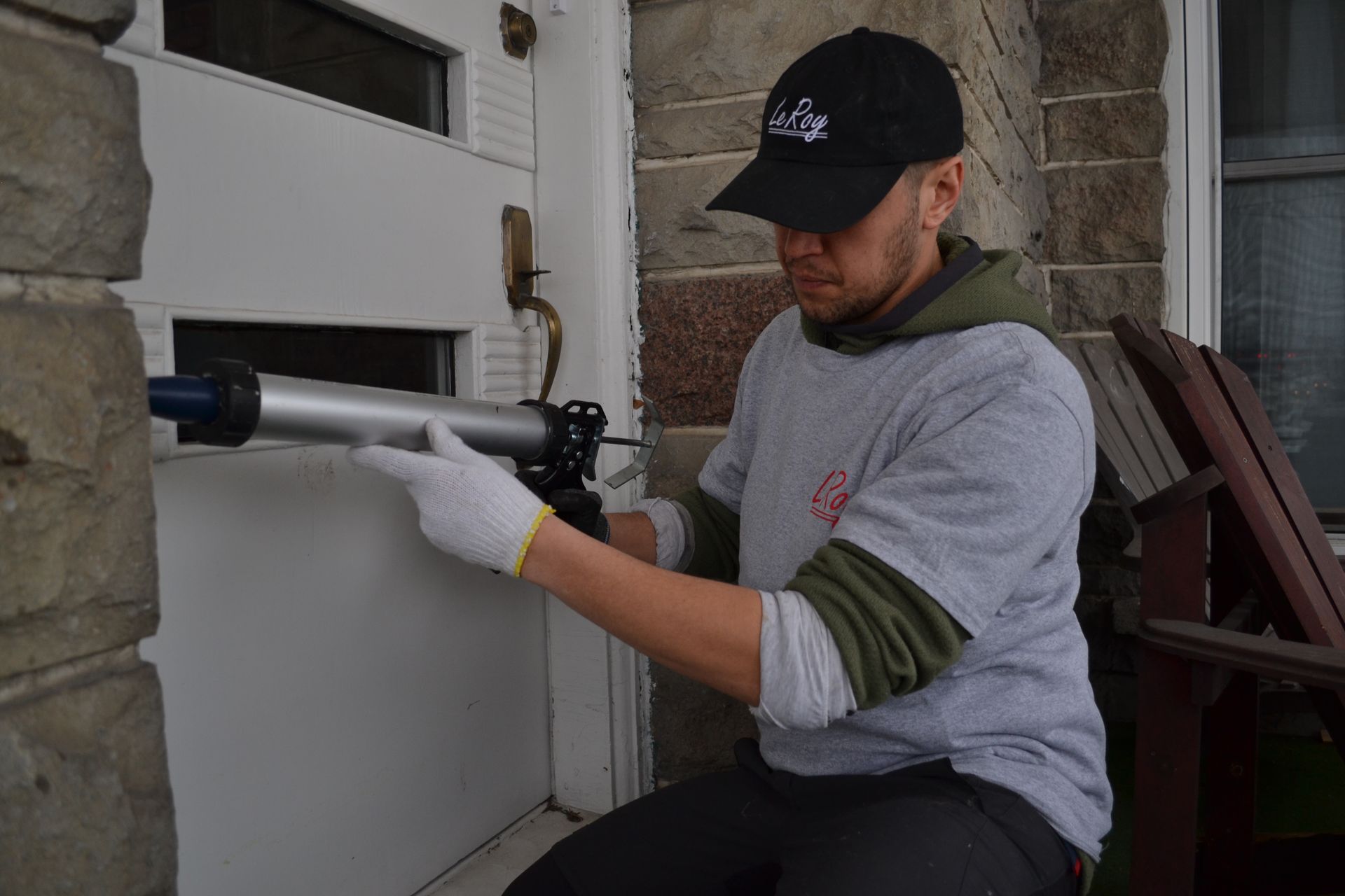 Un homme portant une casquette noire et des gants calfeutrant un cadre de porte blanc sur une maison en pierre.