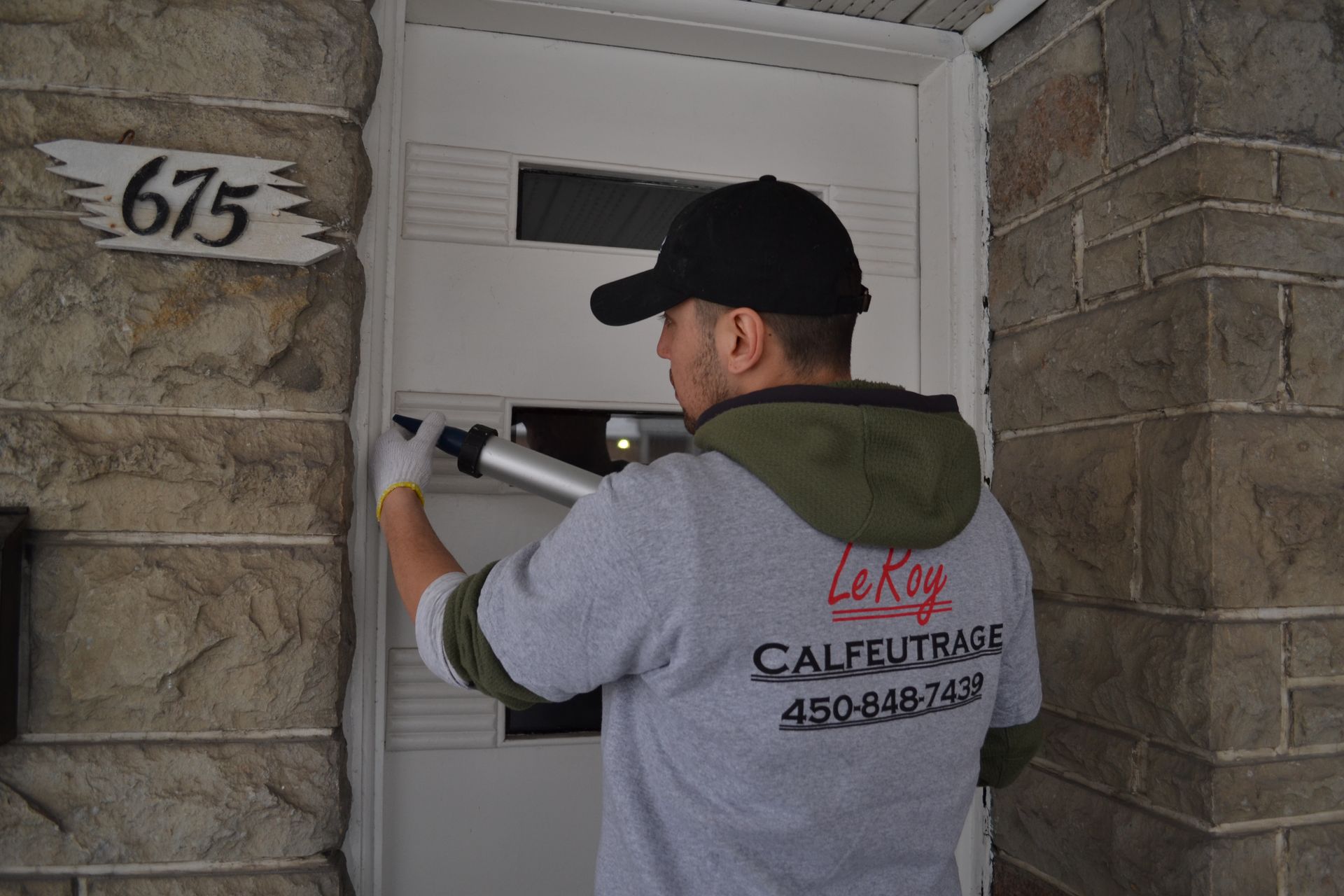 Un homme calfeutre le cadre d'une porte blanche sur un bâtiment en briques. Le numéro 675 est inscrit au-dessus de la porte.