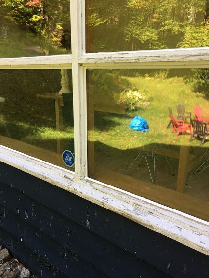 Une fenêtre à cadre blanc reflète une cour herbeuse avec des chaises rouges et un objet bleu ; autocollant ADT sur le coin du cadre.