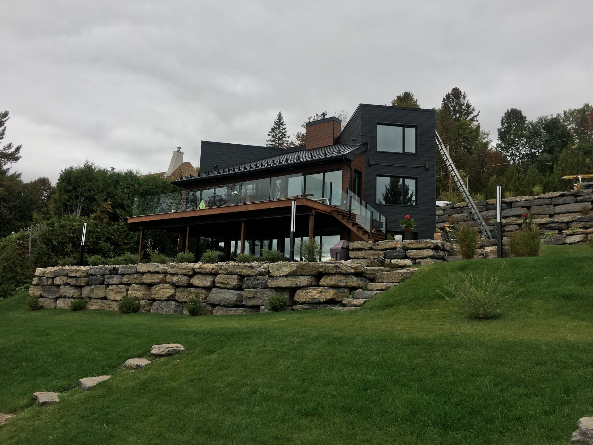 Maison moderne avec extérieur sombre, grandes fenêtres et aménagement paysager en pierre à plusieurs niveaux sur une colline herbeuse.