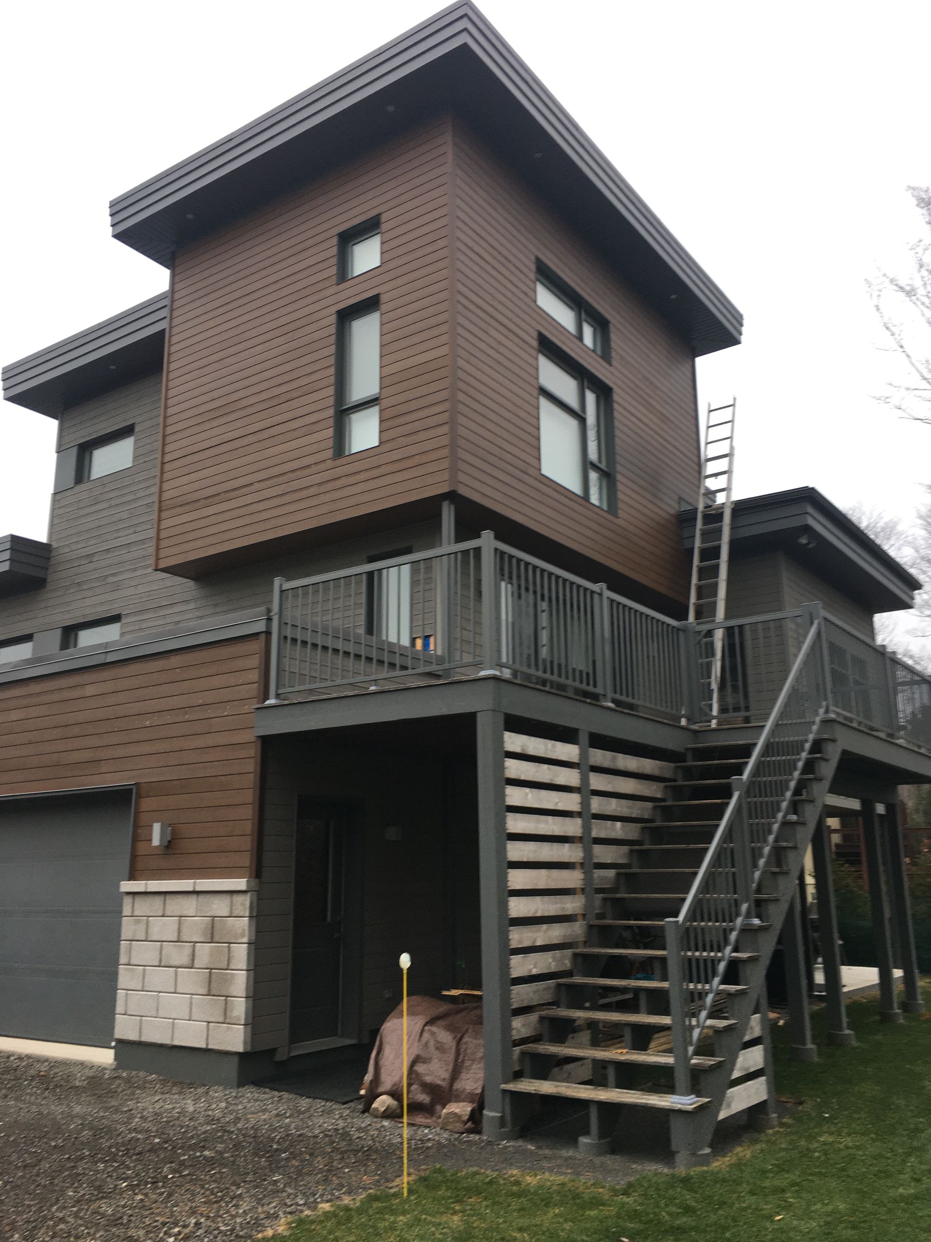 Maison moderne à deux étages avec revêtement brun et gris, terrasse et escalier. Une échelle est appuyée contre le côté.
