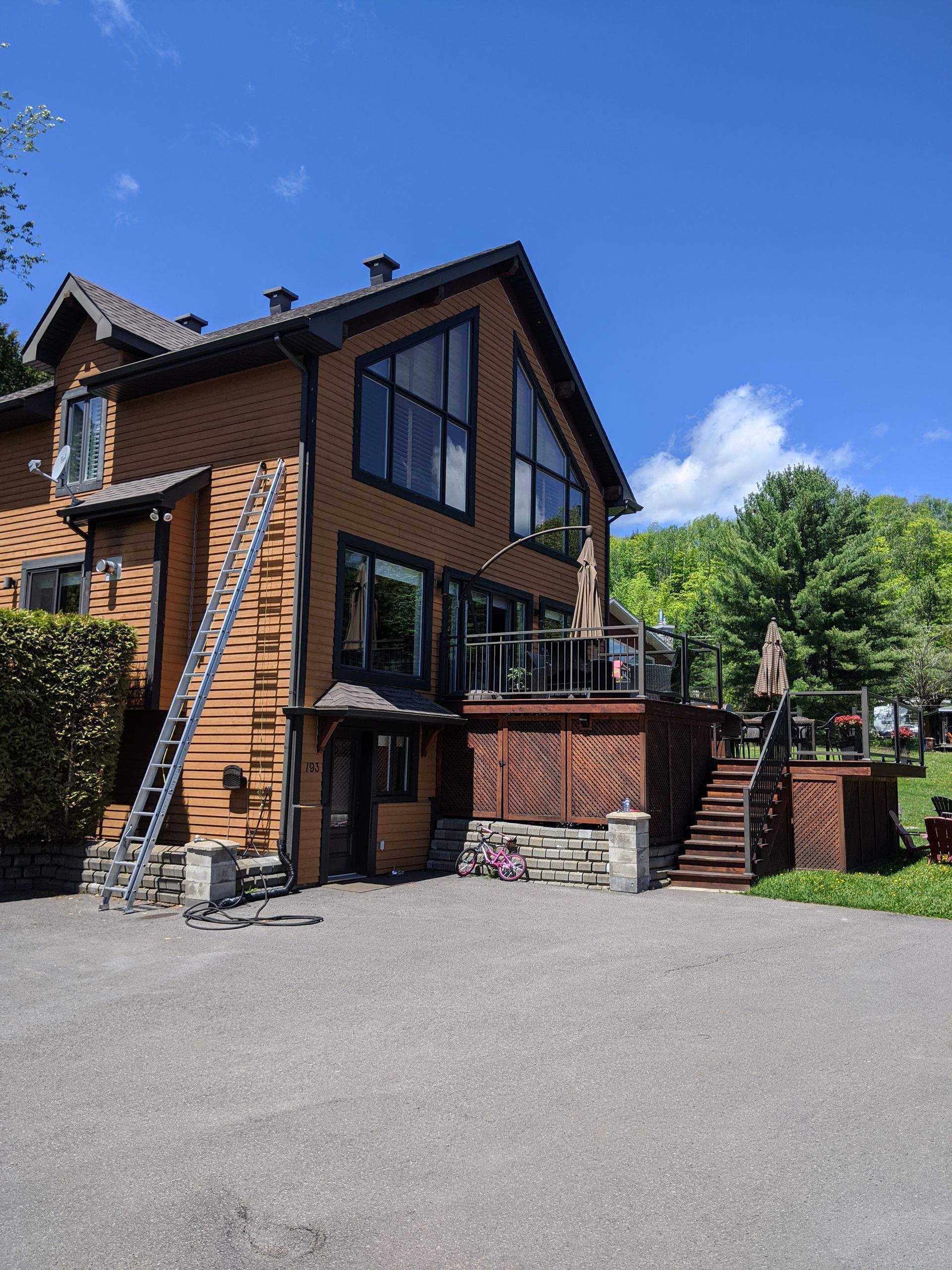 Maison brune à deux étages avec des garnitures sombres et de grandes fenêtres, accessible par une allée de gravier et des escaliers en bois.