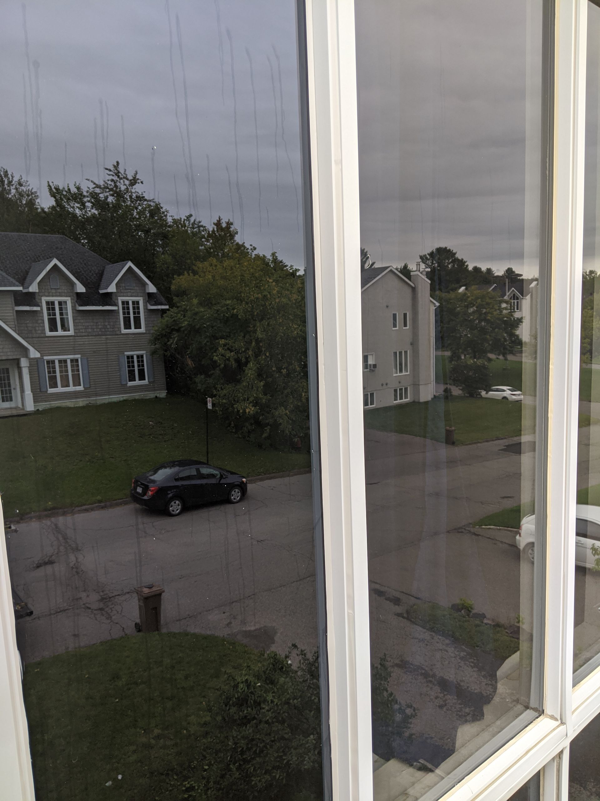Vue depuis une fenêtre d'une rue avec une maison, une voiture et un immeuble. Ciel couvert.