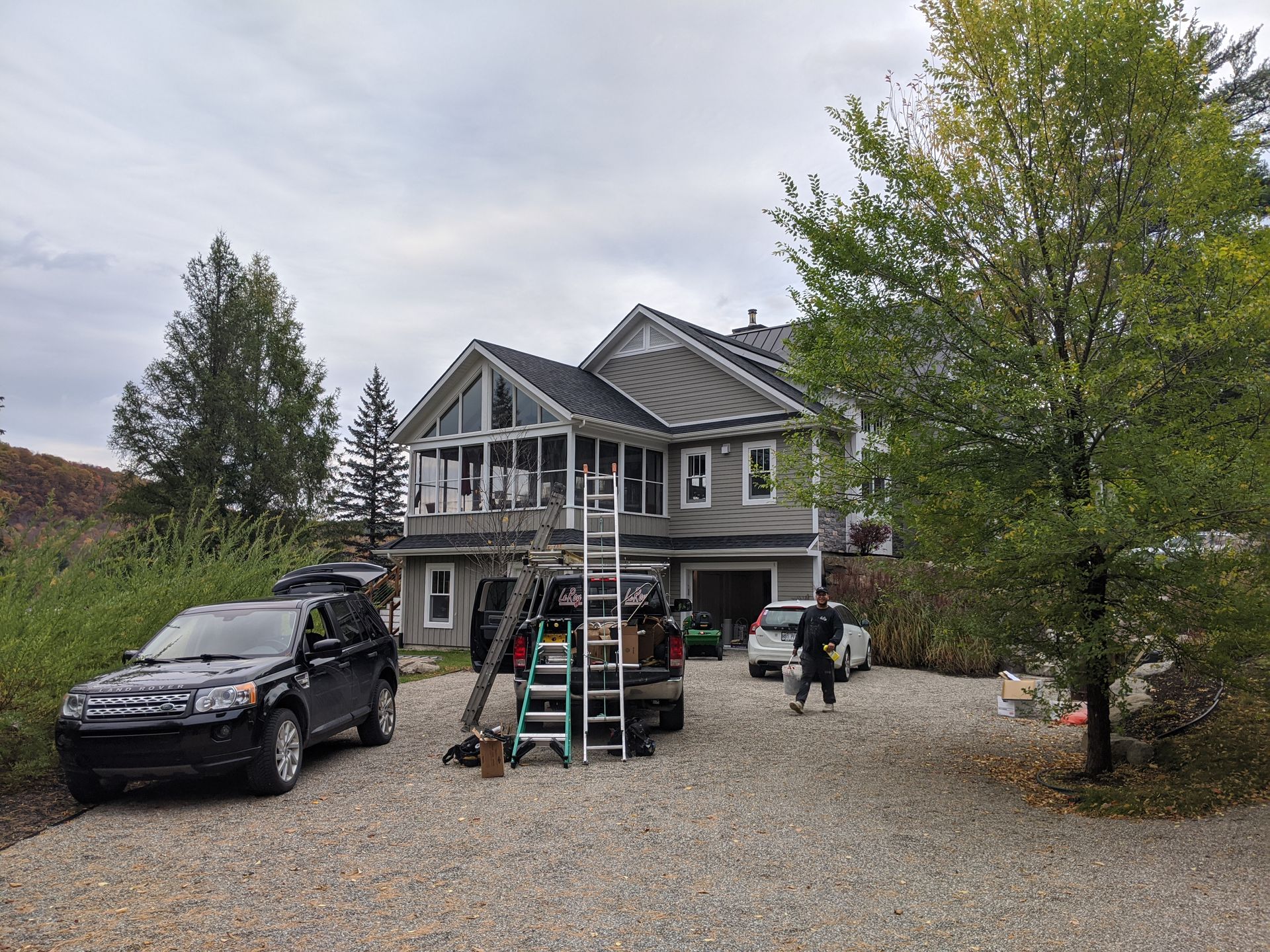 Maison à deux étages avec revêtement gris et toit foncé ; les voitures et les ouvriers sont dans l'allée de gravier.