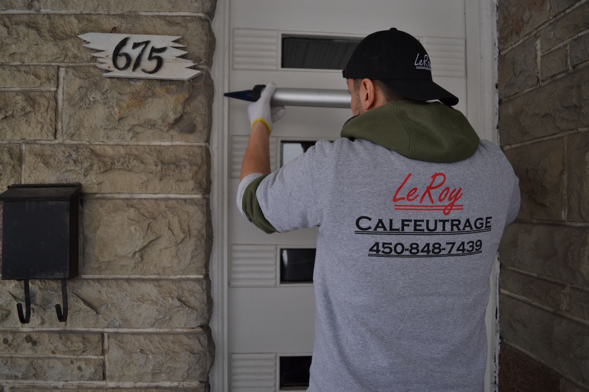 Un homme calfeutre une porte d'entrée blanche, portant une chemise avec « Le Roy Calfeutrage » et un numéro de téléphone dessus.