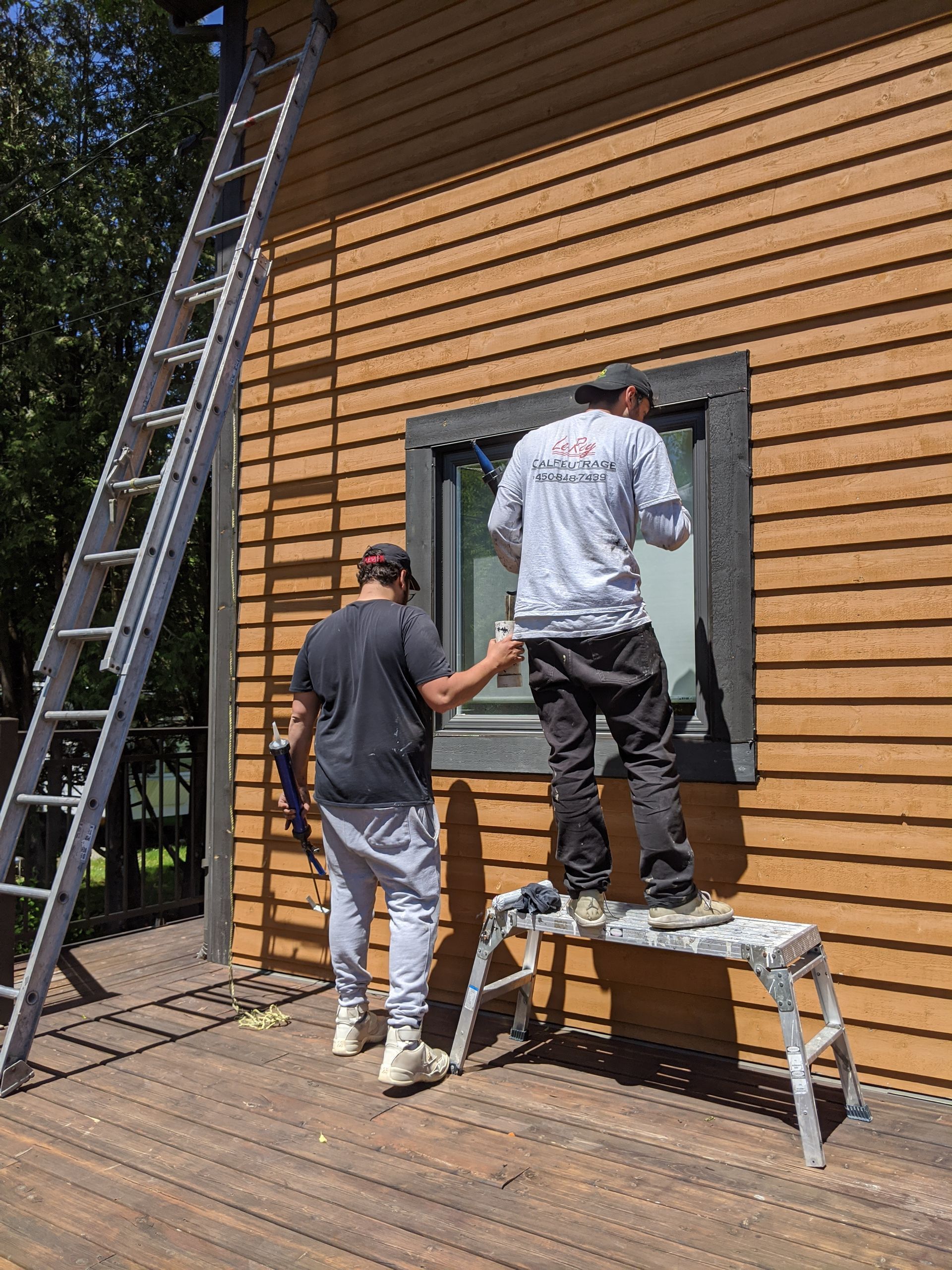 Deux hommes peignent un cadre de fenêtre sur une maison brune ; l'un sur un escabeau, l'autre tenant un pot de peinture et une échelle.