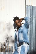 Man in denim jacket and pants, hand on head, leaning against a textured wall.