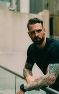 Man with tattoos on arms, leaning on a railing, wearing a black shirt, looking away from the camera.