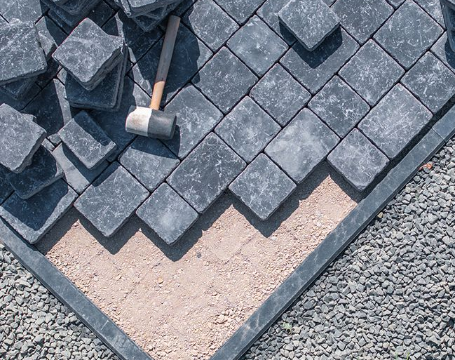 Installation of dark gray pavers on a sand base, with gravel border and mallet.