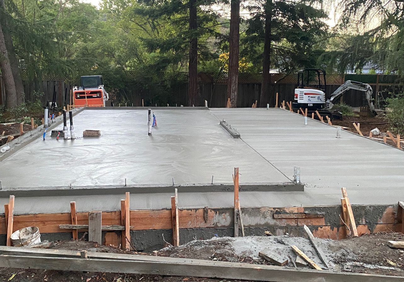 Freshly poured concrete foundation for a structure; construction site with equipment and wooden framing in a wooded area.