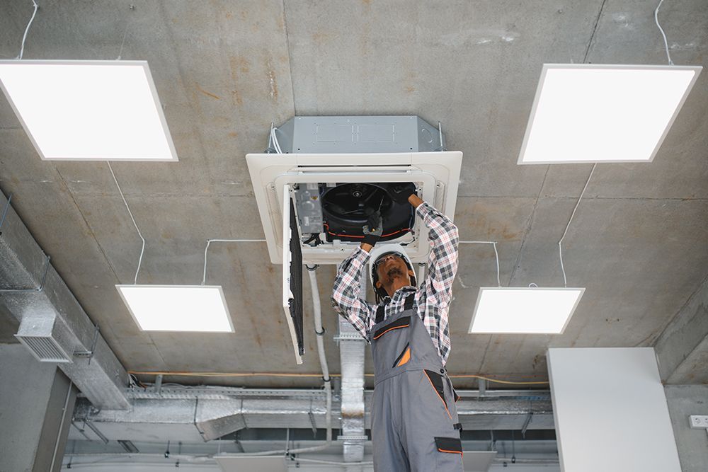 A technician in a plaid shirt and overalls repairs a ceiling-mounted air conditioning unit in a modern office space.