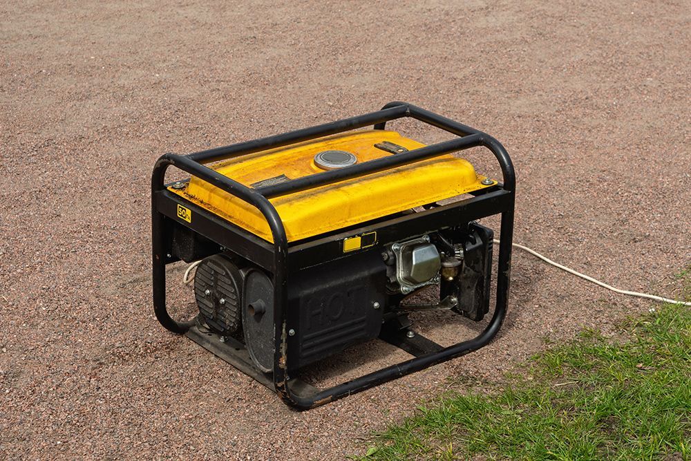 A yellow portable power generator sits on a gravel surface next to a grassy area.