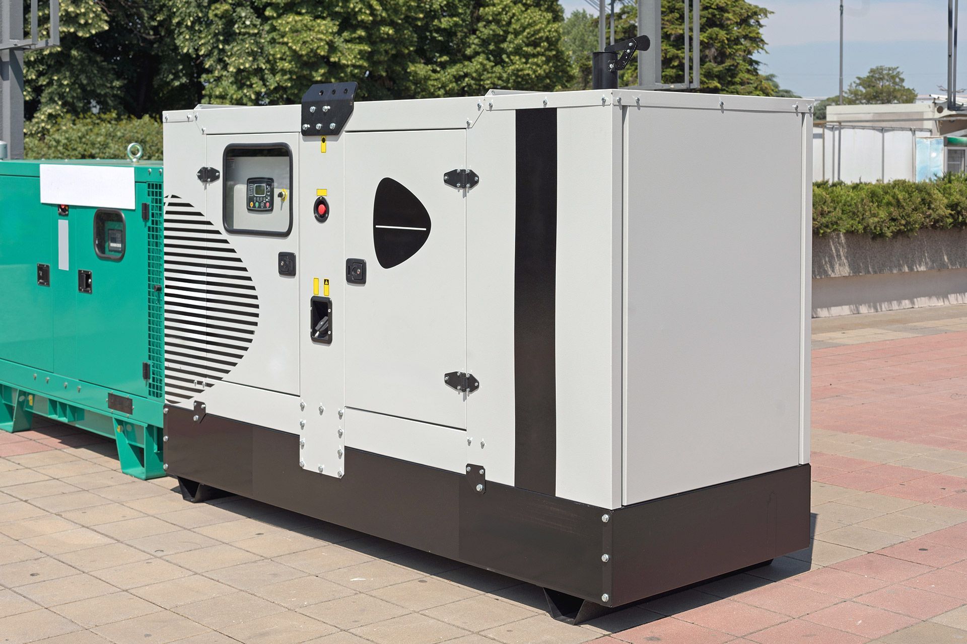 A white and dark-brown industrial power generator standing outdoors on a paved surface next to a green unit.