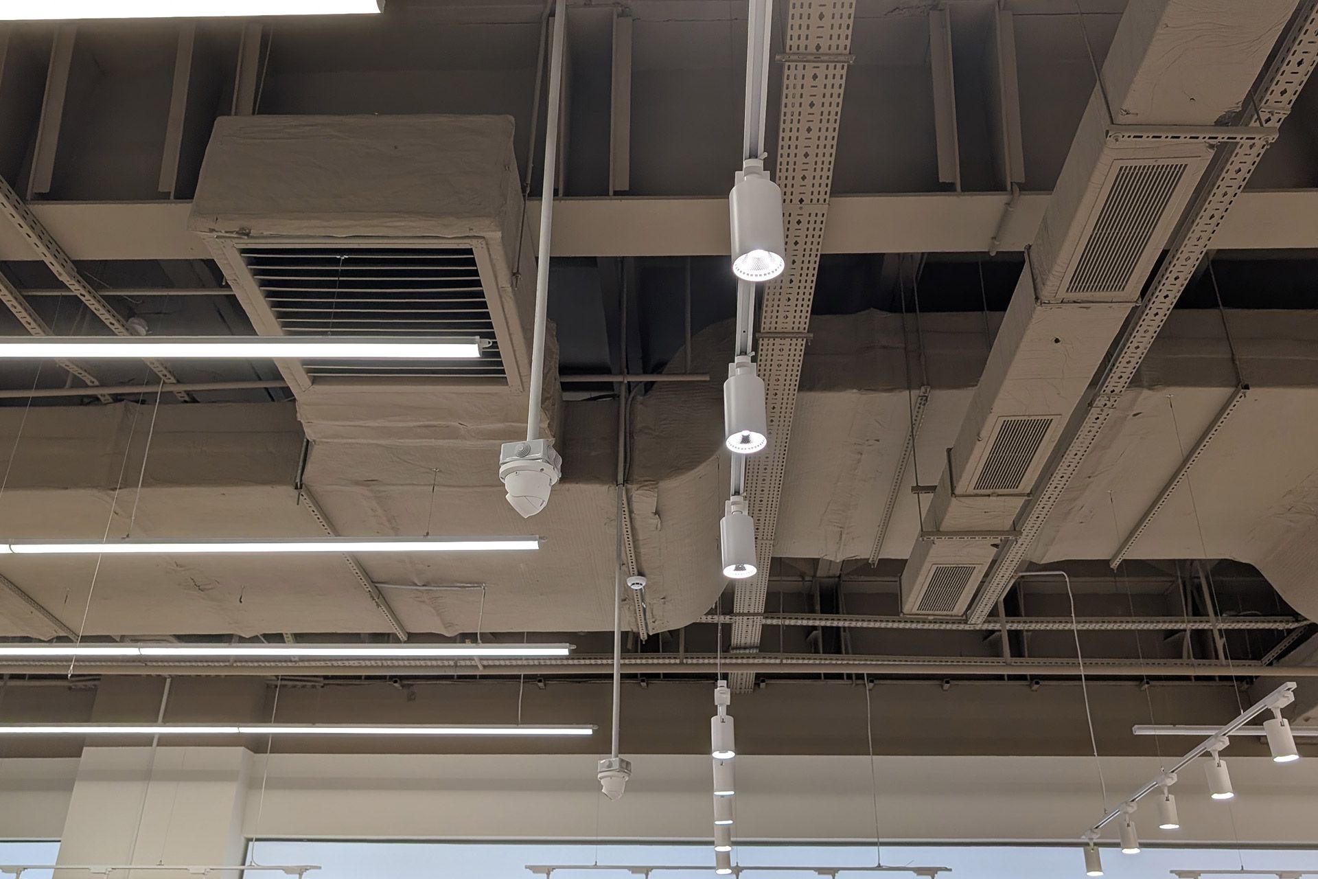 A high ceiling with exposed beige ducts, long linear light fixtures, and a series of white cylindrical pendant lights.