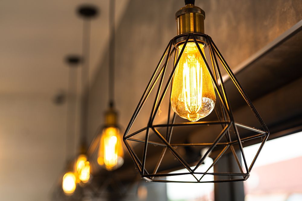 A close-up of a warm, glowing Edison bulb inside a geometric wire cage pendant light, with more blurred lights behind it.