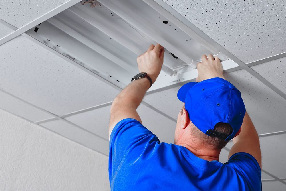 A person in a blue shirt and cap reaching up to repair or install a fluorescent light fixture in a drop ceiling.