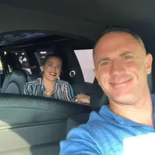 Two smiling people inside a car, one taking a selfie in the front seat and one in the back seat.