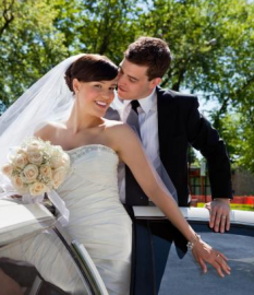 Newlyweds in wedding attire smiling beside a car outdoors, with the groom leaning in affectionately.