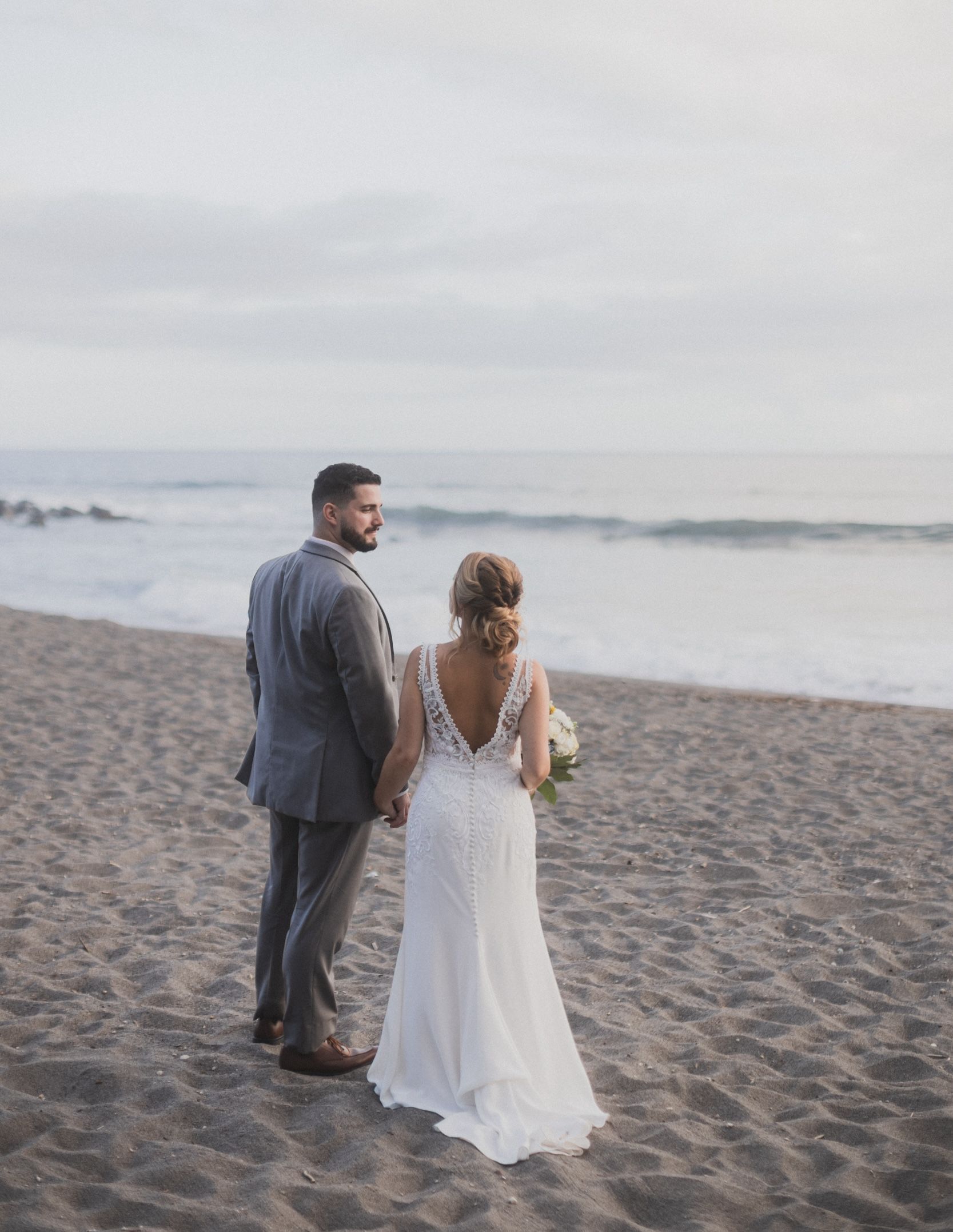A bride and groom are standing on the beach looking at the ocean.