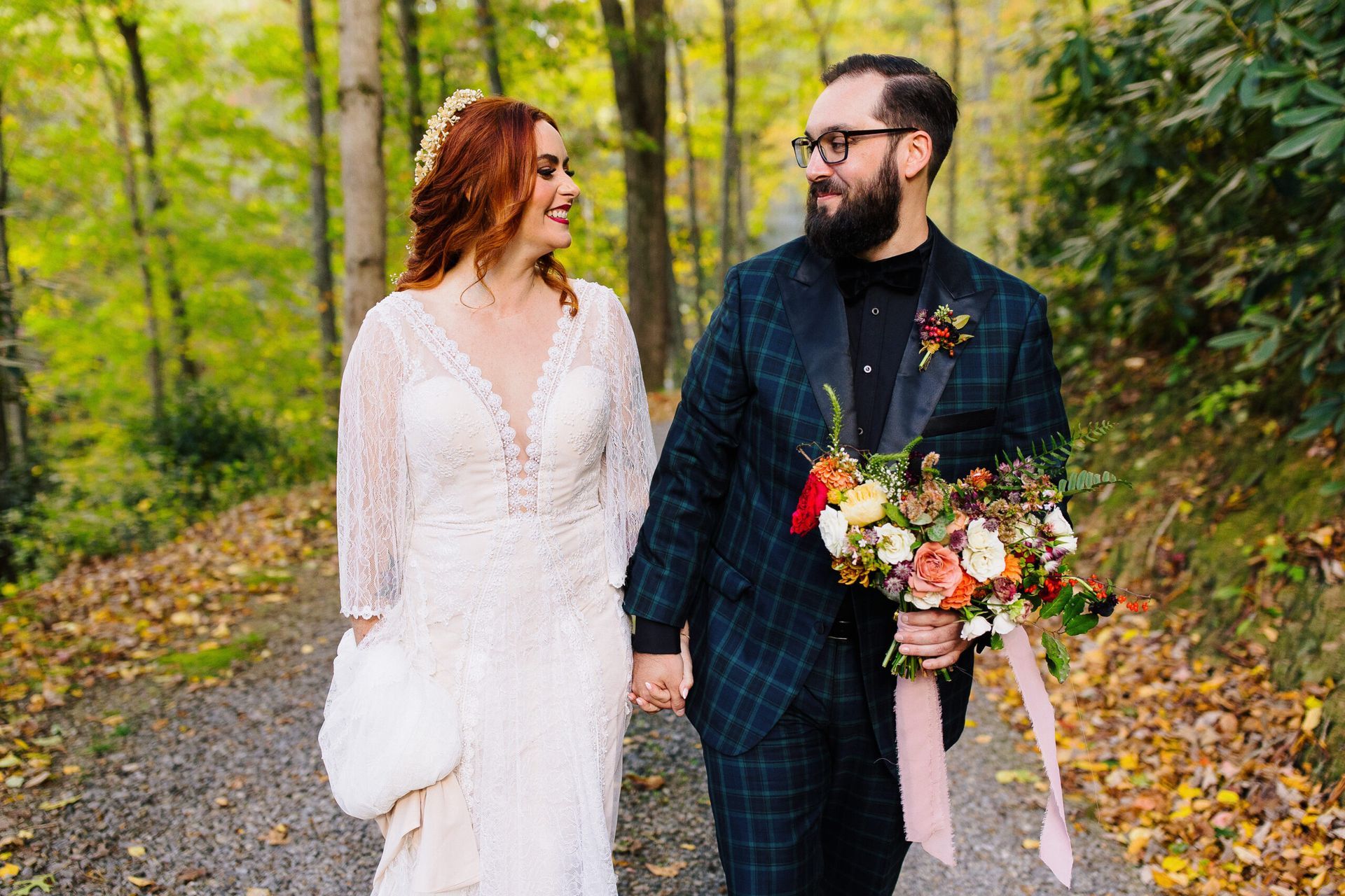 A bride and groom are walking down a path in the woods holding hands.
