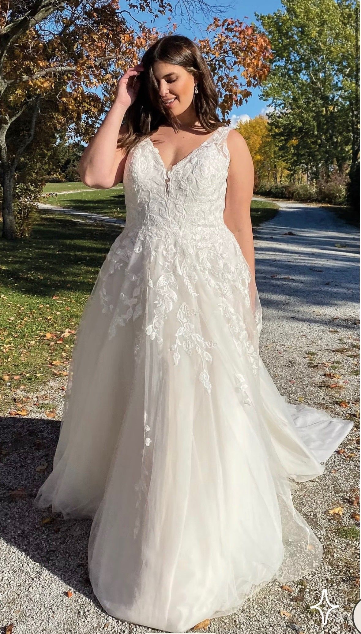 Woman in a white lace wedding dress standing outdoors on a gravel path with autumn trees.