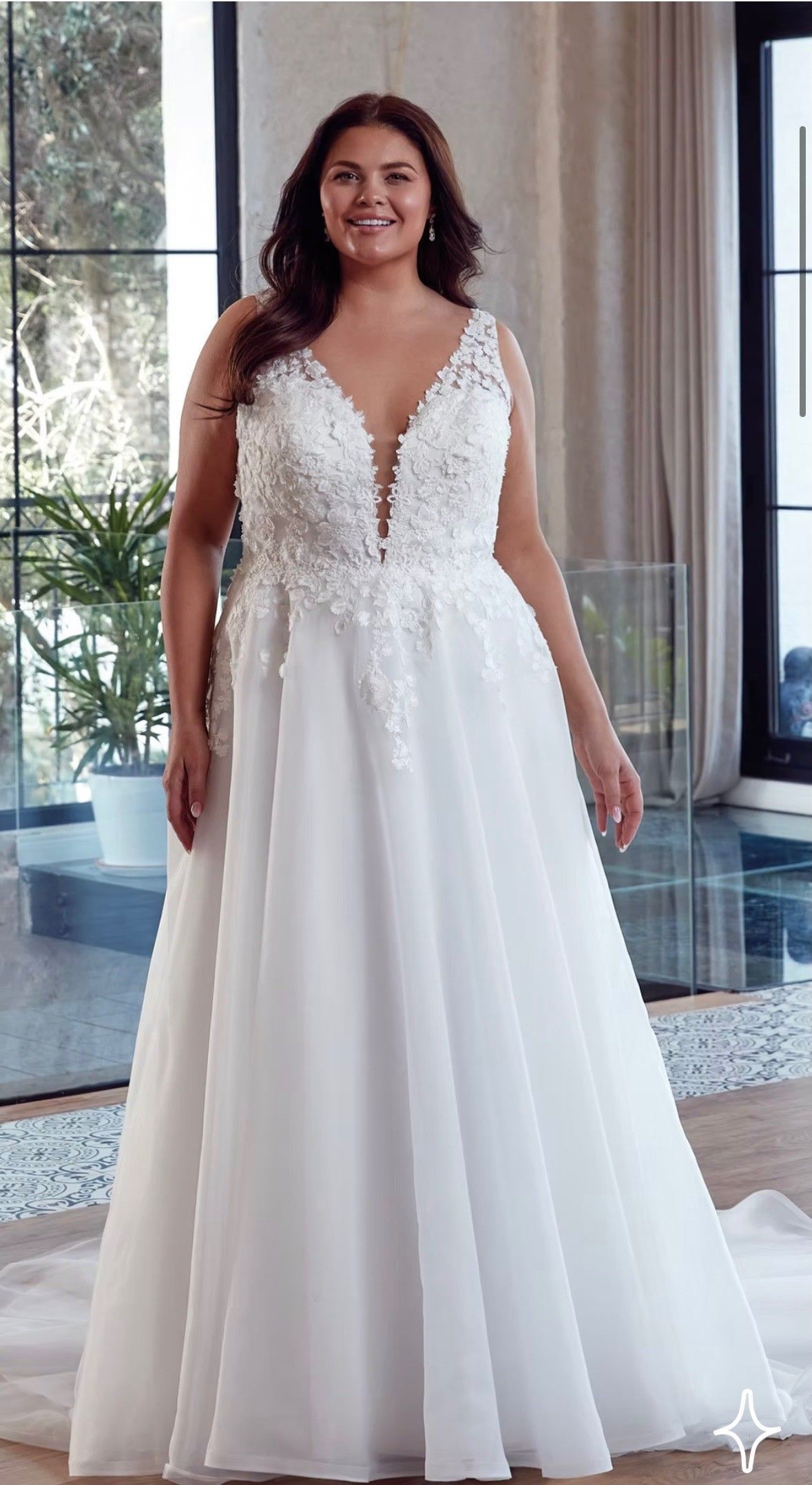 Woman in a white lace wedding dress standing indoors near large windows.