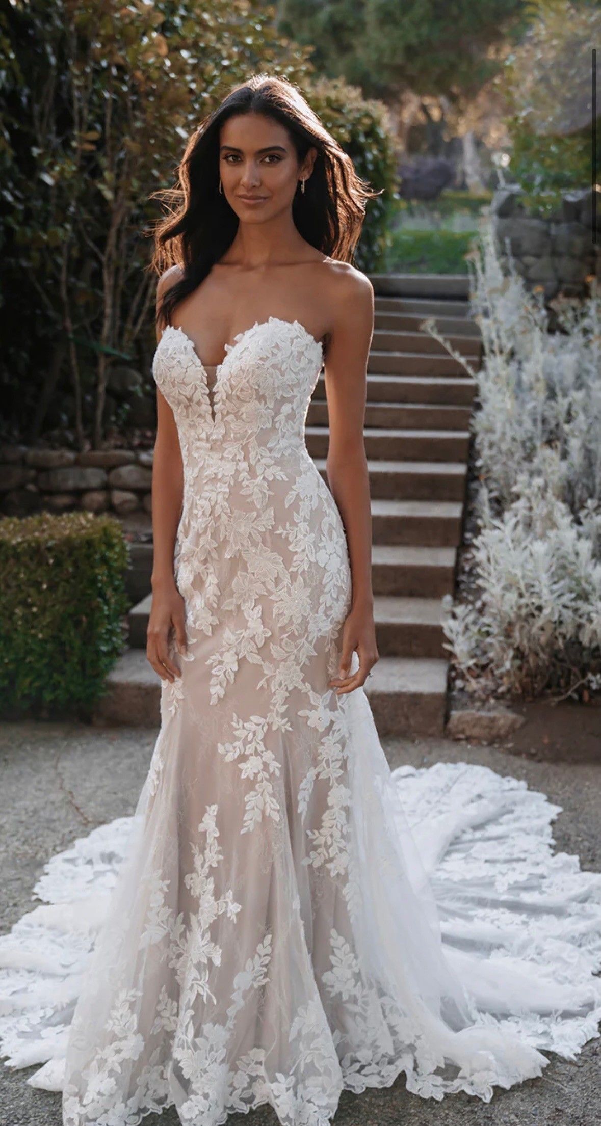 Woman in a white lace wedding gown standing on outdoor stone steps with a long train.