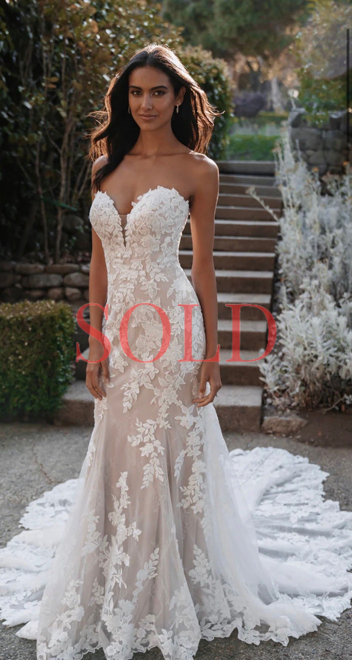 Woman in a white lace wedding gown standing on outdoor stone steps with a long train.