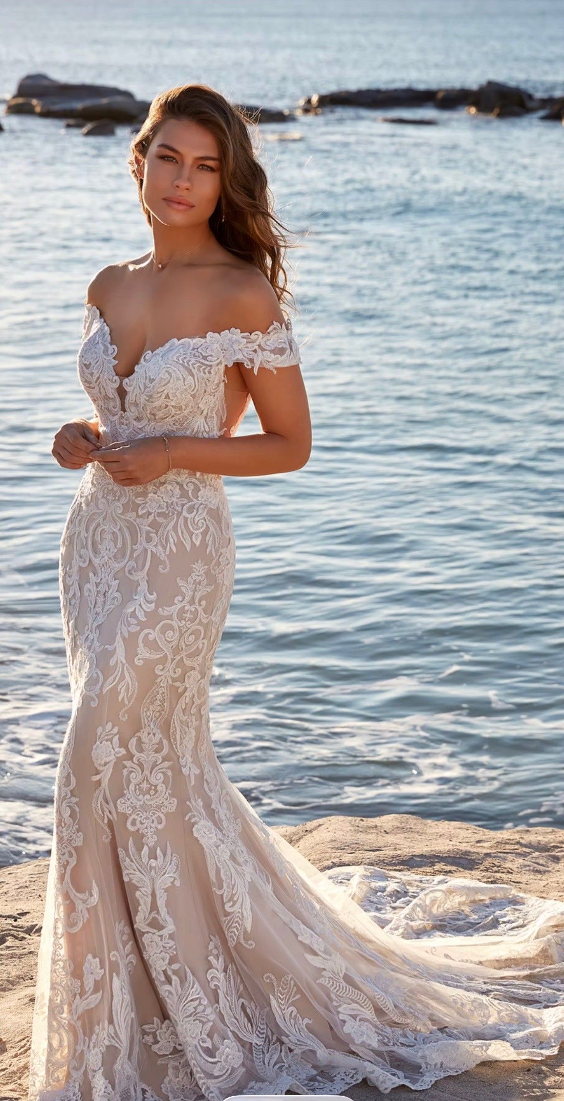 Woman in a white lace wedding dress standing on a beach at sunset.