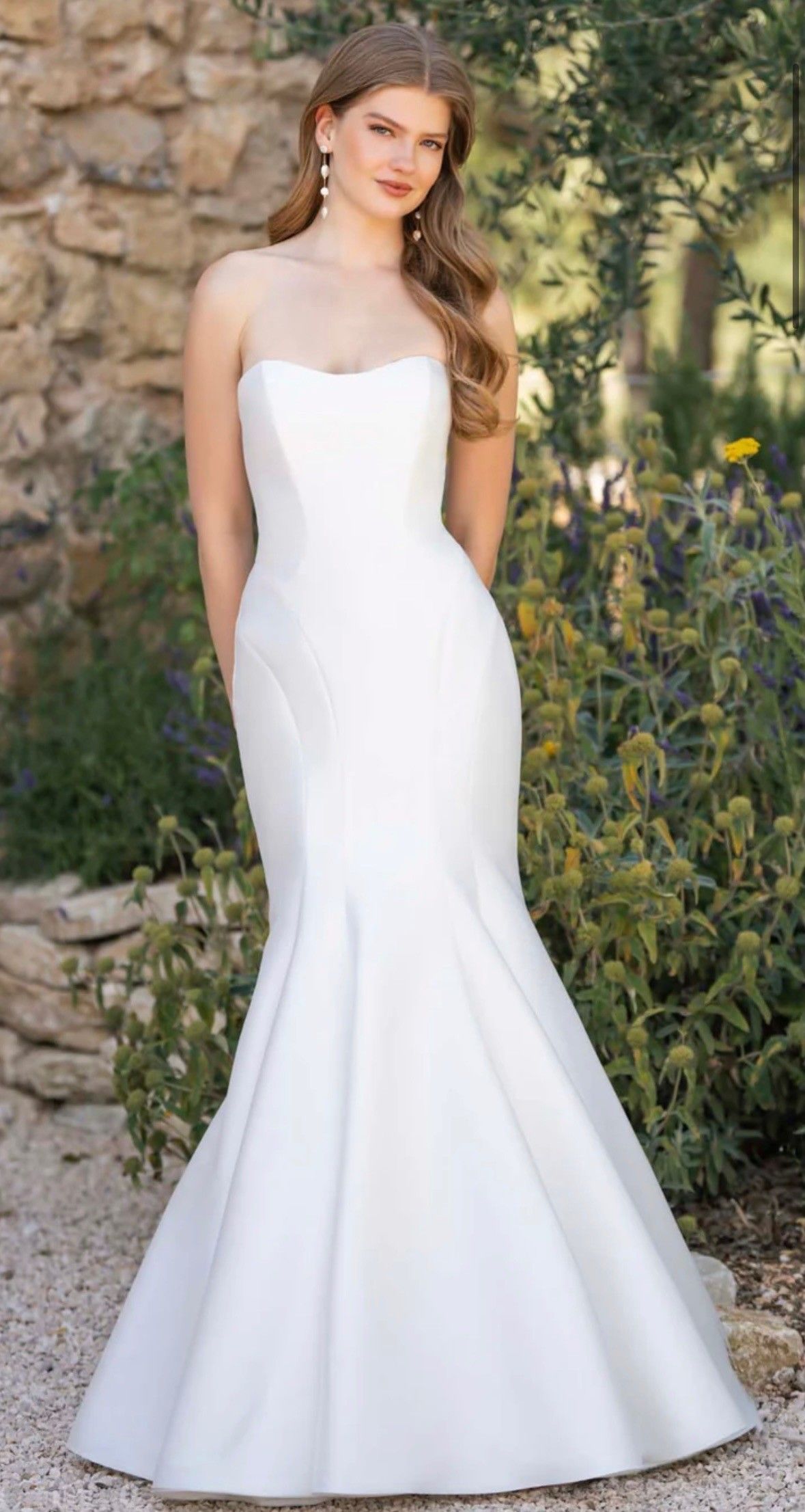 A person standing outdoors wearing a white, floor-length lace wedding dress with a deep V-neckline.